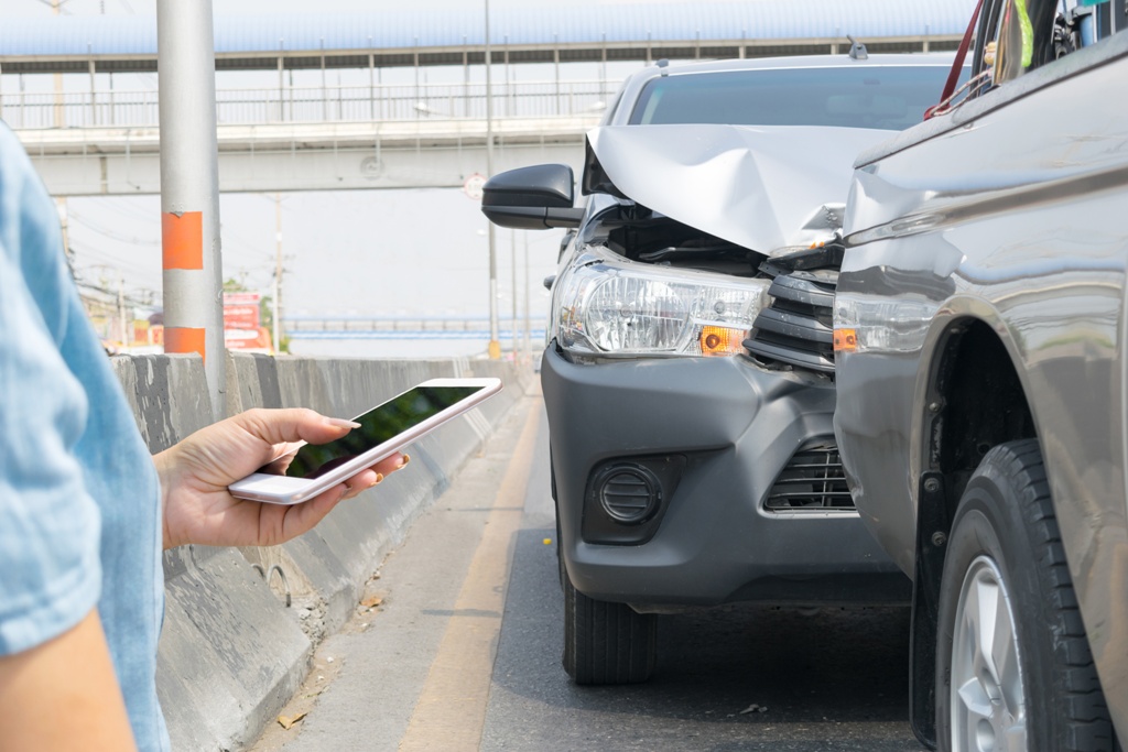 View of a head-on collision between two cars on a road as a woman stands nearby typing on her phone - a stressful situation that a Grab or Gojek loan can help alleviate