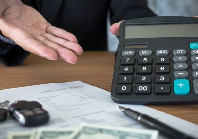 Executive in black suit shows a calculator above a contract, pen, and car keys