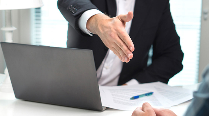 Man in black suit extends his hand for a handshake to seal a fast cash loan deal above a laptop and documents on a desk